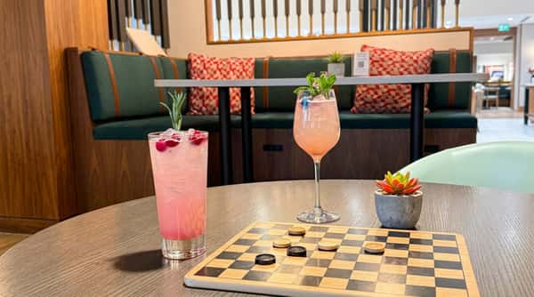 Two pink cocktails with garnishes on a table with a checkers board in a restaurant setting.