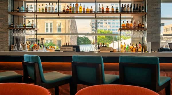Stylish bar interior with teal bar stools and orange chairs in the foreground, looking out to city buildings.