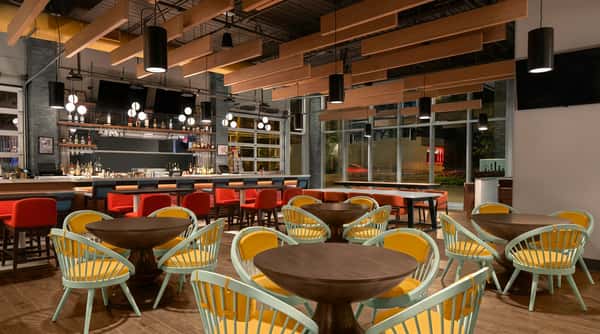 A modern bar and dining area with a long bar, red stools, mint green chairs with yellow cushions, and wood tables.