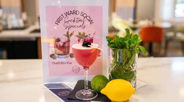 Pink cocktail garnished with a blackberry, next to a pitcher of mint, lime, and lemon, with a "Cocktail Specials" sign in the background.