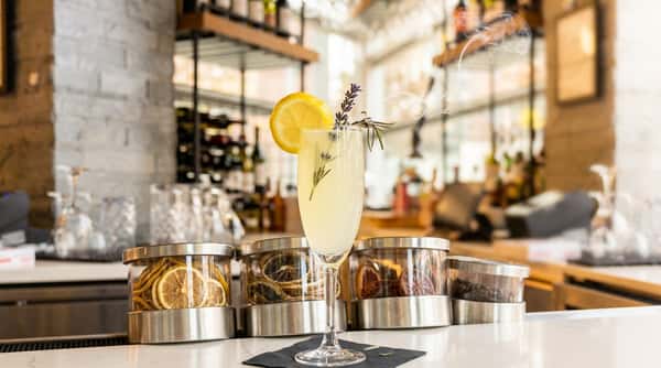 Lemon-colored drink in a tall glass, garnished with a lemon slice and rosemary, emitting smoke.
