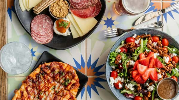 Charcuterie board with meats, cheeses, mustard, and crackers, BBQ flatbread, and a salad with strawberries, nuts, and feta.
