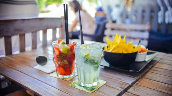 Two cocktails, one strawberry and one lime mojito, with a bowl of yellow chips and salsa on a wooden table.