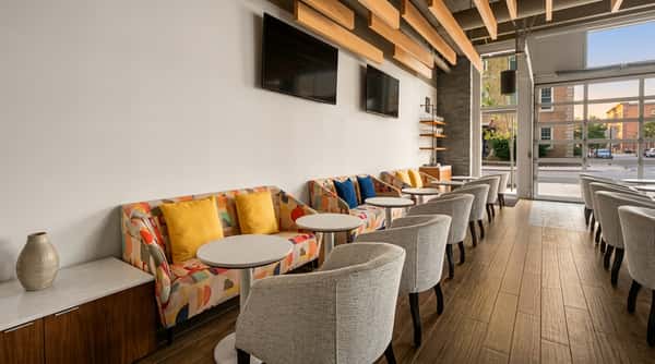 Modern lounge area with patterned sofas and gray armchairs, small white tables, TVs, and wood flooring.