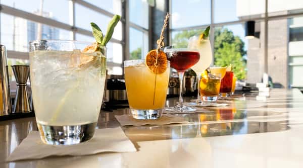 Assorted cocktails on a bar surface, including a clear drink with sage, and a yellow drink with a dried orange slice and lavender sprig.