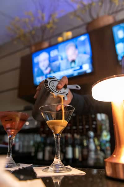 Bartender pouring a freshly mixed cocktail through a metal strainer into a martini glass at Pasta Lovers Trattoria in Midtown Manhattan, NYC, with colorful cocktails and a softly lit bar in the background.