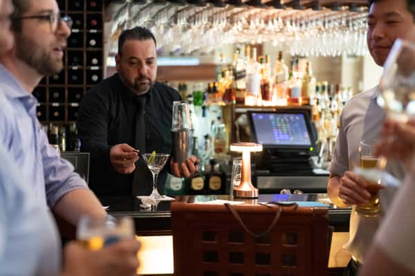 Bartender preparing cocktails behind the bar at Pasta Lovers Trattoria in Midtown Manhattan, NYC, surrounded by wine glasses, bottles, and warm restaurant lighting.