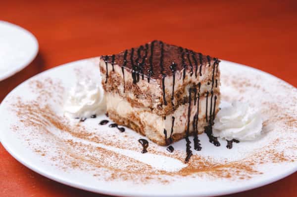 Slice of creamy Italian dessert topped with chocolate drizzle and powdered sugar, served on a white plate with whipped cream at Pasta Lovers Trattoria in Midtown Manhattan, NYC.