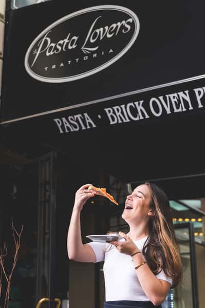 Woman enjoying a slice of Italian pizza outside Pasta Lovers Trattoria in Midtown Manhattan, NYC, beneath the restaurant's Pasta and Brick Oven sign.