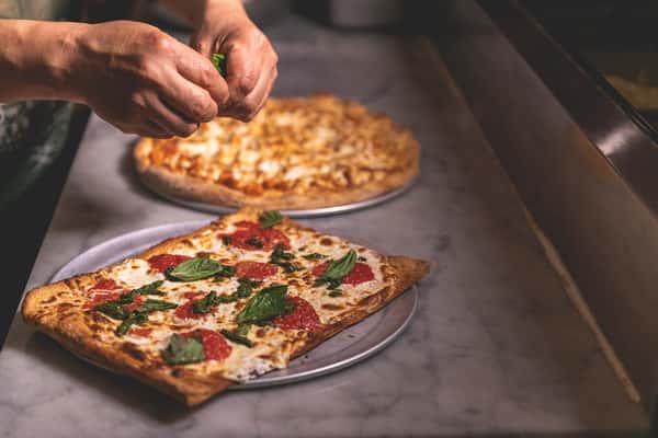 Freshly baked rectangular Margherita pizza topped with tomato sauce, melted mozzarella, and basil, served on a metal tray while another pizza is prepared in the background at Pasta Lovers Trattoria in Midtown Manhattan, NYC.
