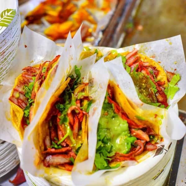 Three tacos al pastor loaded with marinated pork, fresh cilantro, and green salsa, served on yellow corn tortillas with paper wrappers.