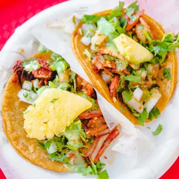 Two tacos filled with al pastor pork, chopped onions, cilantro, creamy green sauce, and pineapple chunks on a white plate.