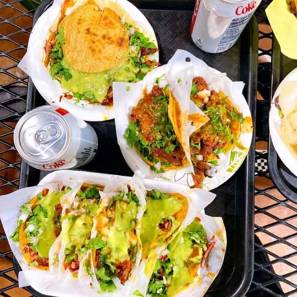 Assorted street tacos topped with green salsa, onions, and cilantro, served with tostadas and Diet Coke cans on a black tray.