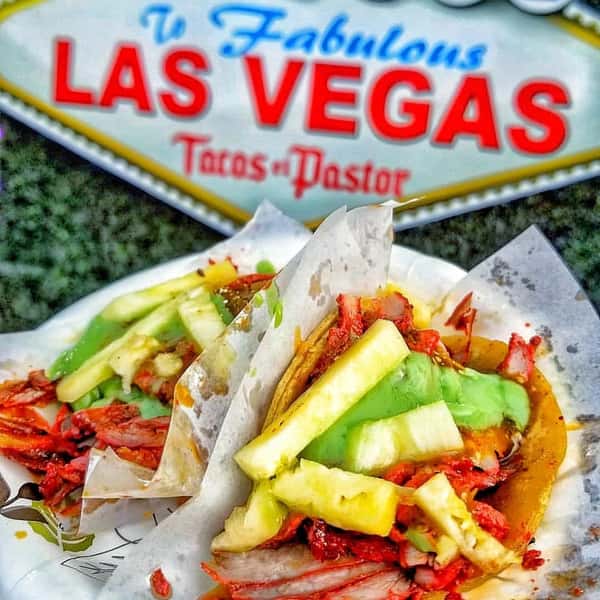 Tacos al pastor topped with pineapple and green salsa, served on paper plates in front of a vibrant Las Vegas sign backdrop.