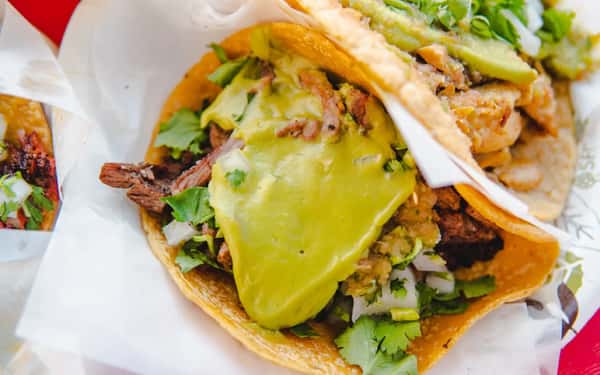 Taco filled with grilled beef, diced onions, cilantro, guacamole, and green salsa on a corn tortilla with another taco partially visible.
