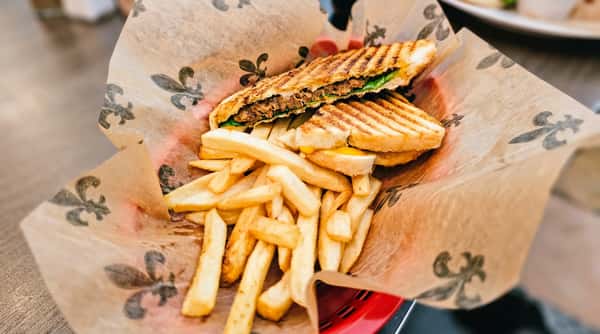 Grilled panini sandwich with seasoned meat, melted cheese, and fresh spinach served with crispy French fries in a paper-lined basket at a casual café.