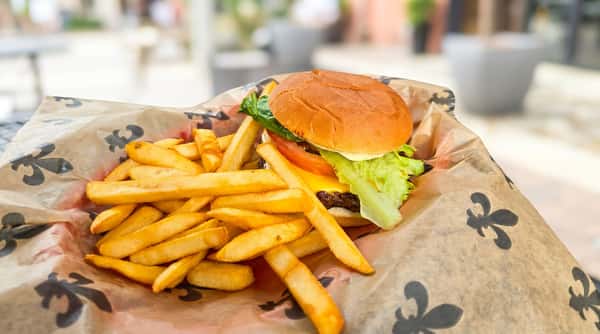 Cheeseburger with fresh lettuce, tomato, pickles, and melted cheese served with crispy French fries on a paper-lined basket in a casual outdoor dining setting.