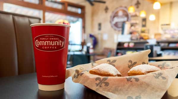 Hot coffee in a Community Coffee cup served with powdered sugar beignets inside a cozy café with warm lighting and inviting seating.