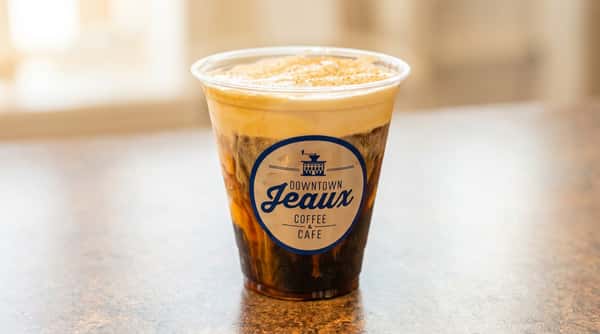 Layered iced coffee topped with cold foam in a Downtown Jeaux Coffee & Café cup, served on a tabletop with a softly blurred café background.
