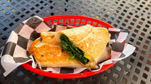 Grilled breakfast wrap with eggs, melted cheese, and fresh spinach, lightly toasted and served in a basket on an outdoor café table.