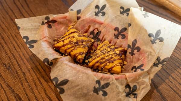 Golden fried beignets drizzled with chocolate sauce and topped with cookie crumbles, served in a paper-lined basket on a rustic café table.