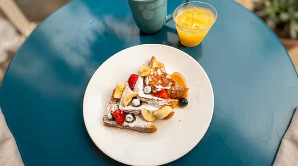 French toast topped with fresh strawberries, bananas, blueberries, powdered sugar, and syrup, served with orange juice and coffee on an outdoor café table.