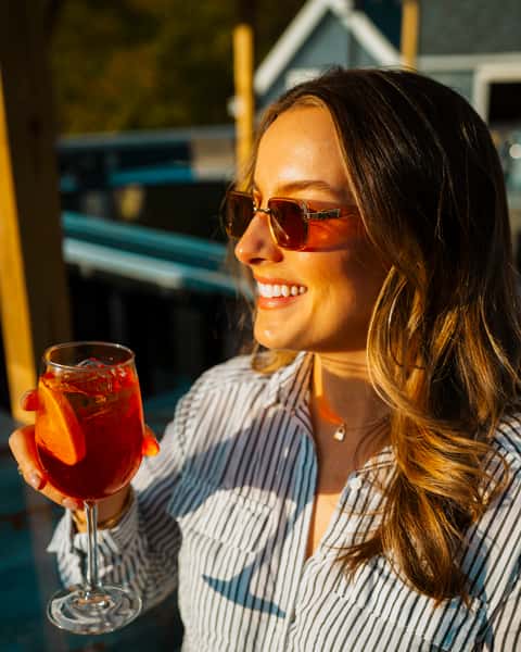 Smiling woman enjoying a fruity cocktail garnished with citrus at an outdoor patio during golden hour — a relaxed and vibrant setting perfect for rooftop bars or beachside restaurants.