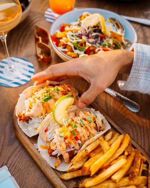 Hand reaching for fish tacos topped with slaw, diced peppers, and creamy sauce, served with golden fries and a fresh lime wedge — a flavorful coastal dish ideal for seafood eateries or casual outdoor dining.