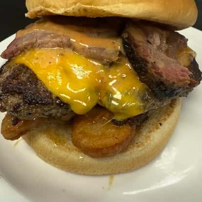 A close-up of a cheeseburger with cheddar cheese, grilled beef patty, and smoked brisket slices topped with sauce, all in a sandwich bun.