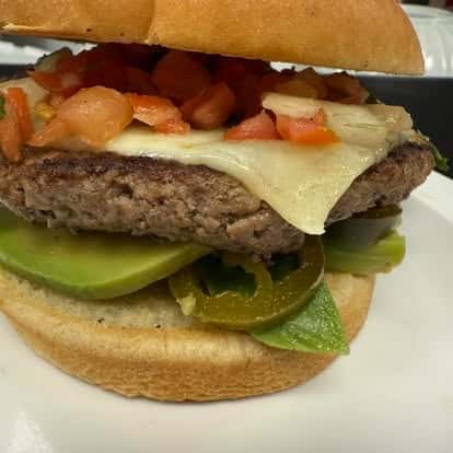 A freshly made burger with melted cheese, tomatoes, avocado, jalapeños, and leafy greens, all stacked in a soft bun.