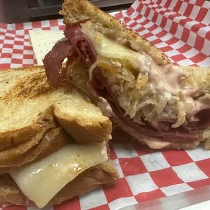 A grilled sandwich with melted cheese and corned beef, featuring sauerkraut spilling from the sandwich, served on a red-checkered paper.