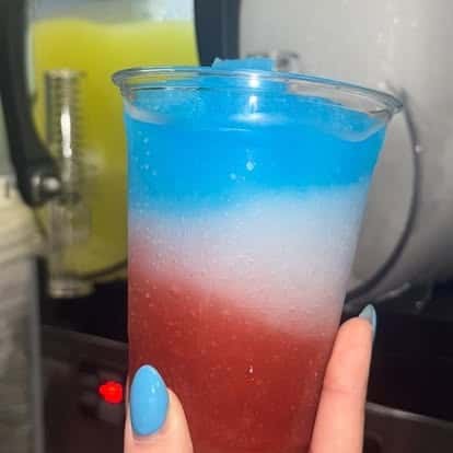 A layered drink in red, white, and blue, held in a clear cup with the blue layer at the top, resembling a patriotic slushie.