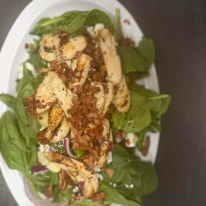 A spinach salad topped with grilled chicken, crumbled bacon, and red onions.