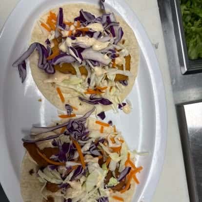 Two tacos filled with fried fish, topped with shredded purple cabbage, shredded white cabbage, and orange carrots, drizzled with sauce.