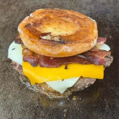 A close-up of a burger featuring a beef patty, melted cheese, crispy bacon, and a glazed donut bun.
