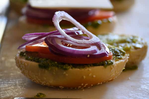 Close-up of a sesame bagel topped with pesto, sliced tomatoes, and red onions in a café kitchen