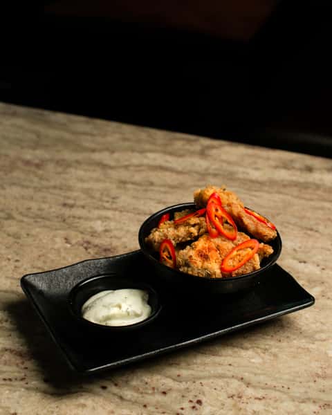Crispy fried chicken bites topped with sliced red chilies, served with creamy dipping sauce on a sleek black plate in a contemporary Asian restaurant