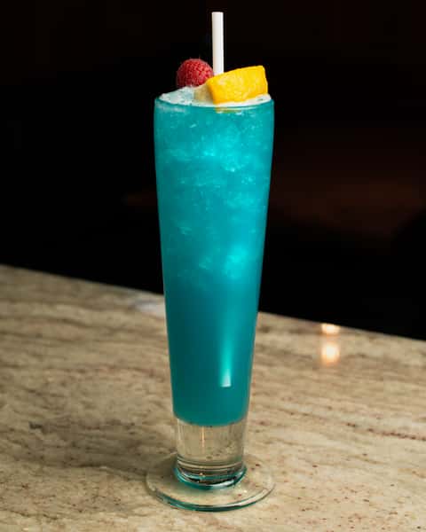 Bright blue tropical cocktail garnished with raspberry and citrus, served in a tall glass on a marble bar in a stylish nightlife setting