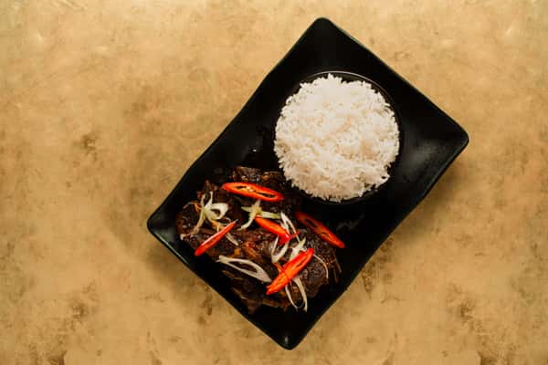Overhead view of tender braised beef with chili garnish and fluffy white rice, plated on a sleek black dish in a contemporary Asian restaurant