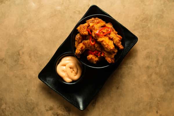 Crispy fried chicken bites topped with sliced red chilies, served with creamy dipping sauce on a sleek black plate in a modern Asian restaurant setting