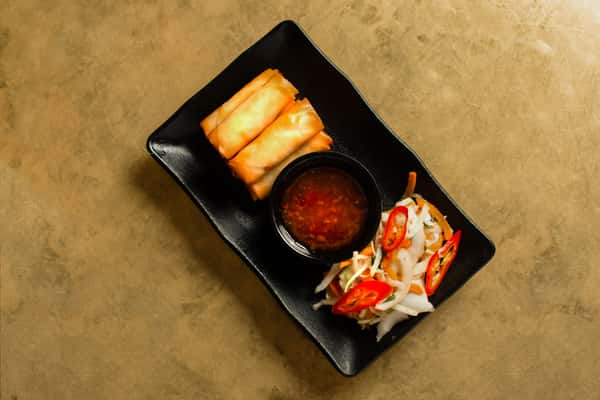 Crispy golden spring rolls served with sweet chili dipping sauce and fresh Asian slaw with sliced red chilies, presented on a modern black plate in a stylish restaurant setting