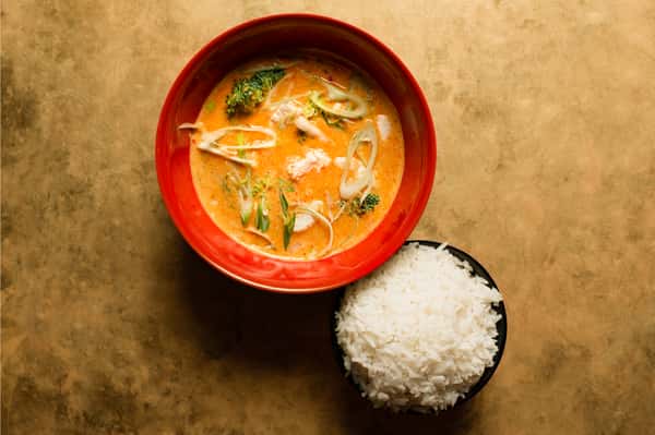 Creamy coconut curry soup with fresh vegetables and herbs, served in a vibrant red bowl alongside a side of steamed white rice in a modern Asian restaurant setting