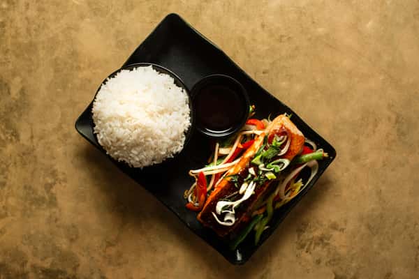 Grilled Asian-style salmon fillet with mixed vegetables and savory sauce, served with steamed white rice on a modern black plate