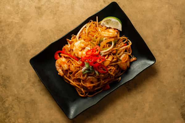 Spicy shrimp noodle stir-fry with vegetables, bean sprouts, and red chilies, garnished with lime on a modern black plate in a vibrant Asian dining setting