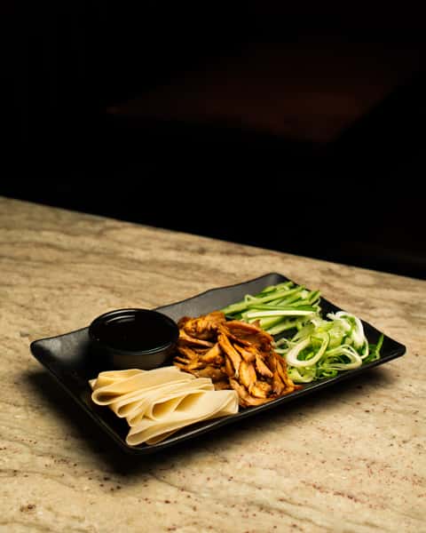 Asian-style duck pancake platter with sliced roasted duck, scallions, cucumber, and hoisin sauce, served on a modern black plate