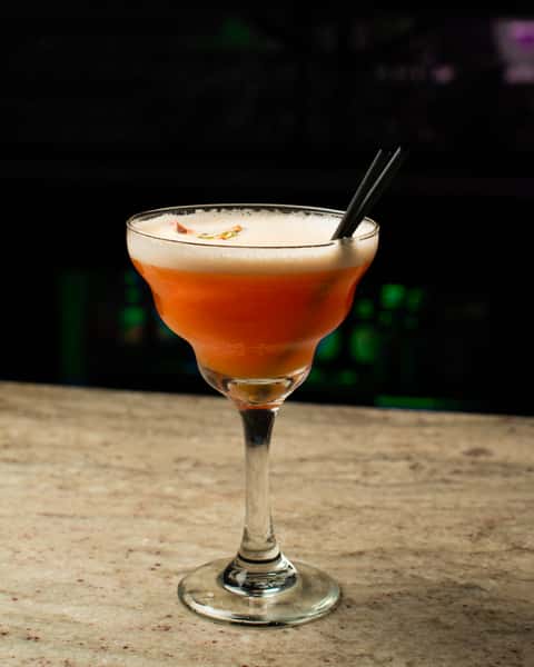 Fruity pink cocktail with frothy top served in a coupe glass, garnished and presented on a marble bar in a modern lounge