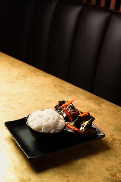 Braised Asian beef short ribs topped with sliced red chilies and scallions, served with steamed white rice on a modern black plate in a stylish restaurant setting