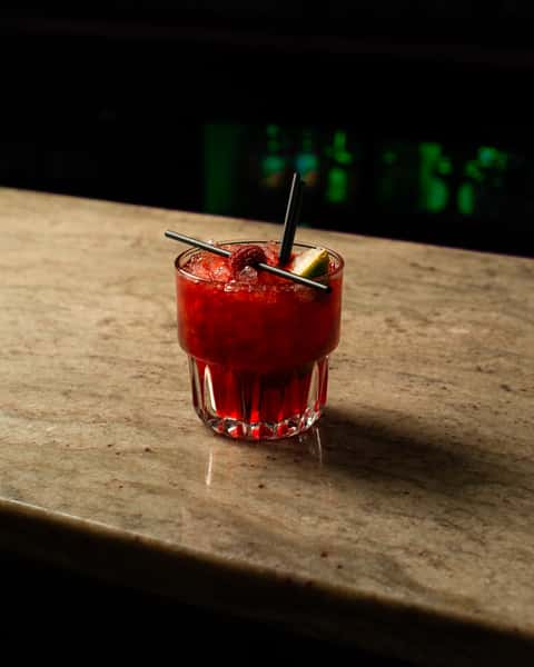 Vibrant red crushed-ice cocktail with lime and raspberry garnish, served in a short glass on a marble bar in a dimly lit bar setting