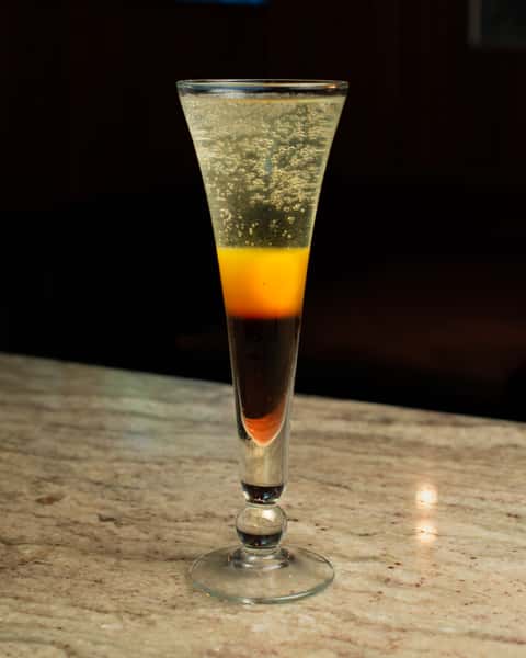 Layered sparkling cocktail with dark, orange, and bubbly top layers in a tall flute glass, served on a marble bar in an upscale lounge setting