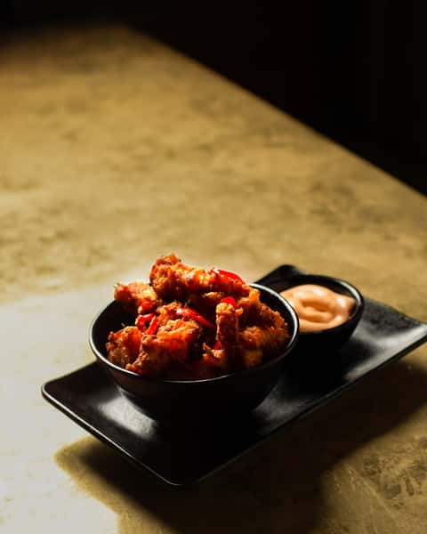 Spicy crispy fried chicken pieces with red chili garnish, served with creamy dipping sauce on a black plate in a dimly lit restaurant